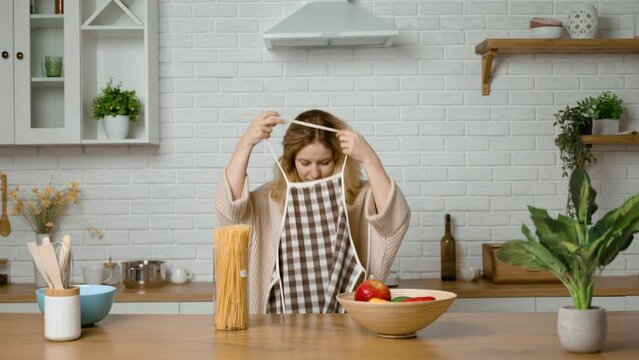 The housewife starts cooking in kitchen and puts on apron. Woman wears cloth that protects her clothes from dirt. The woman loves cooking and enjoys the process of preparing delicious, healthy dishes.
