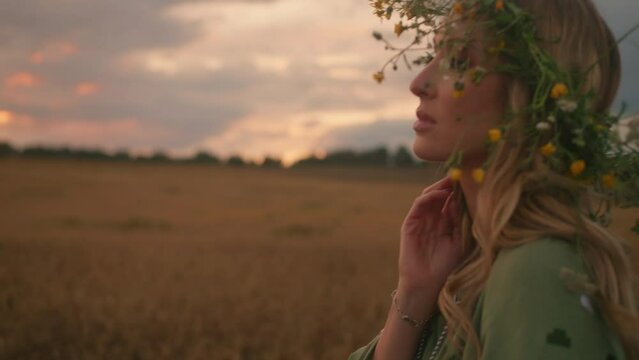 Beautiful young woman with floral crown walks through the wheat field at dawn. A hopefull look into the future, professional bright make up, enigmatic face. Sunset out of city. High quality 4k footage