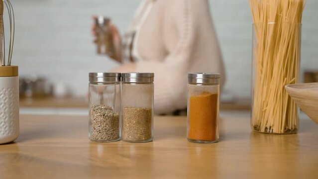 Housewife prepares dinner and takes jar of spices from table. Selection of spices in glass containers. Fragrant and aromatic dry crushed herbs. Spices will add expressiveness and richness of taste 
