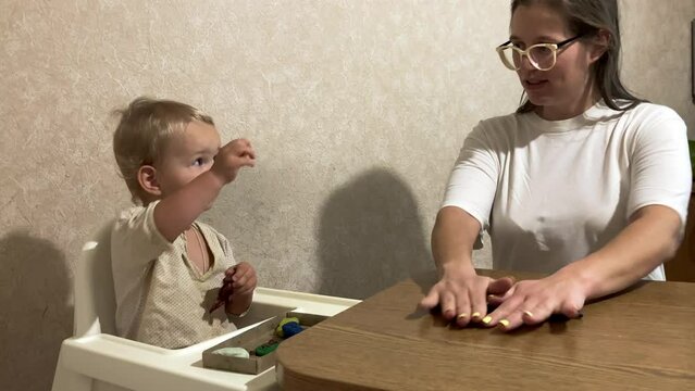 The mother and tha baby are talking. Momming is making the long rope from the modelling clay, when the toddler is choosing the color for the masterpiece. The boy is touching the plasticine.