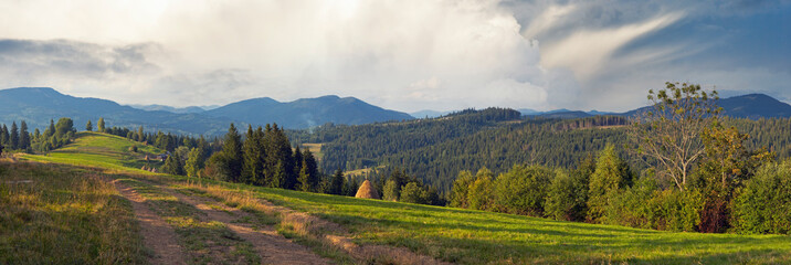 Mountain evening panorama view with earth road, haystackes and country estate (Slavske village outskirts, Carpathian Mts, Ukraine).