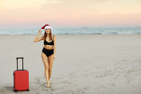 Smiling beautiful young woman in swimsuit, Santa Claus hat on head and red suitcase with mock up on the sand beach with sea background on sunset. Travel concept for Christmas holidays and New Year.