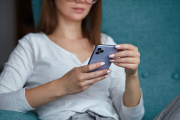 Young woman using mobile phone holding in hands while sitting in armchair at home