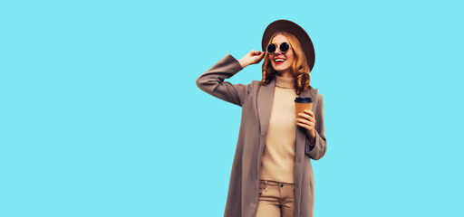 Portrait of stylish beautiful young woman model with cup of coffee wearing brown round hat and coat on blue background