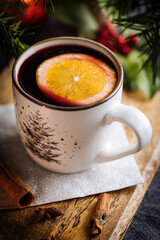Mulled wine with cinnamon and orange served in festive Christmas decoration on wooden board. Party and New Year atmosphere. Christmas tree in background, a mug with Christmas motive. 