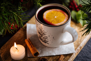 Obraz na płótnie Canvas Mulled wine with cinnamon and orange served in festive Christmas decoration on wooden board. Party and New Year atmosphere. Christmas tree in background, a mug with Christmas motive. 