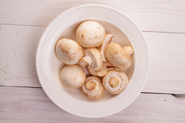 Several organic fresh appetizing mushrooms champignon in a white ceramic plate, close-up, on a table of natural wood, painted white, top view.