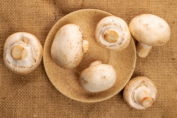 A few whole organic fresh mouth-watering mushrooms champignon, with a ceramic saucer, on a jute napkin, top view.