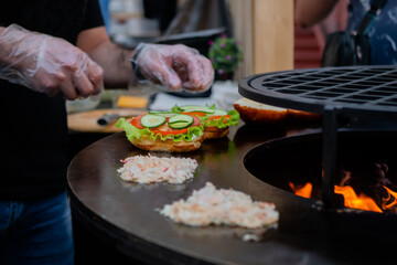 Chef preparing burgers with seafood, fish on brazier at summer local food market - close up view. Outdoor cooking, gastronomy, cookery and street food concept