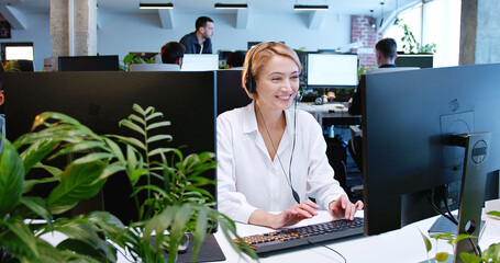Caucasian beautiful woman in headset working at computer screen, talking with client online....