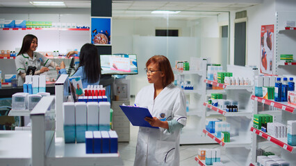 Obraz premium Asian pharmacist working on pills inventory with papers, taking notes about pharmaceutics stock to sell to customers. Drugstore employee looking at boxes of drugs and bottles of vitamins.