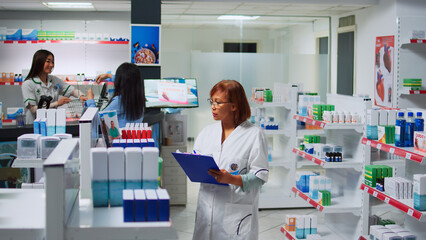 Asian pharmacist working on pills inventory with papers, taking notes about pharmaceutics stock to sell to customers. Drugstore employee looking at boxes of drugs and bottles of vitamins.