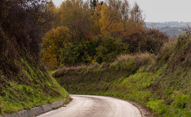 Kręta droga asfaltowa przebiegająca przez wzgórza pogórza Gór Świętokrzyskich .Szczyt jesieni . Drzewa w przepięknych barwach . Na tle szarego , zachmurzonego nieba . 