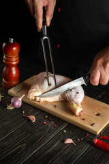 Professional chef or chef cuts a fresh raw chicken drumstick on a dark background. Nearby on the kitchen table are ingredients for cooking and spices