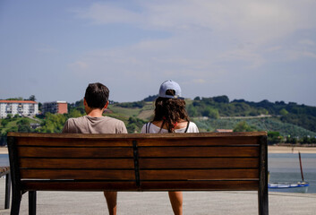 View from Behind of a Heterosexual Couple Sitting on a Bench Observing the Blue Sky and Calm Sea with a Sailboat and Mountains in the Background. Concept of Enjoy Life and Relax