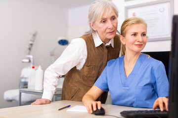 Fototapeta premium Interested elderly female client looking at computer screen while cosmetologist writing examination results and upcoming procedures in aesthetic medicine office