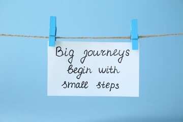 Card with phrase Big Journeys Begin With Small Steps hanging on rope against light blue background....