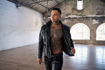 hot tattooed man in leather against the big window.Handsome Young Athletic Male Fashion Model.Muscular athletic sexy male with naked torso. Brutal handsome man with tattooed body.