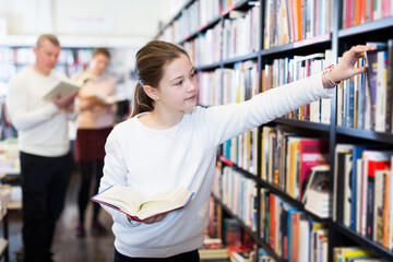 Positive tweenager girl searching for textbooks on bookshelves in store
