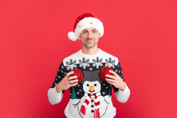 Santa decoration christmas ball. Studio portrait of guy wears santa hat and winter sweater. Man with christmas hat over isolated red background.