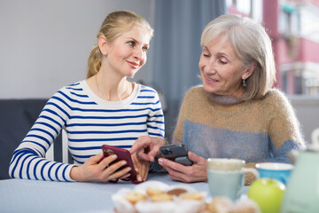 Mature woman with her adult daughter, sitting at a table in a room, looking at photos on a mobile phone