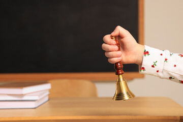 Pupil with school bell near desk and chalkboard in classroom, closeup. Space for text