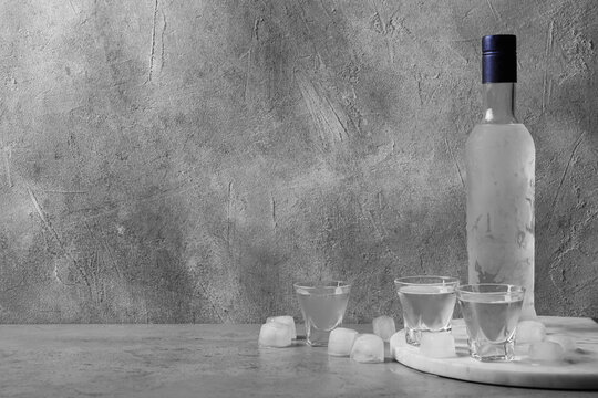 Bottle of vodka and shot glasses with ice cubes on table against grey wall. Space for text