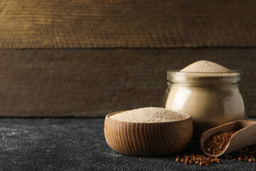 Jar and bowl of buckwheat flour near scoop with grains on black table against wooden wall. Space for text