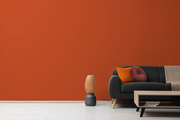 Stylish grey sofa with colorful pillows, wooden table and vase near dark orange wall indoors, space...