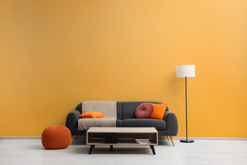 Stylish grey sofa with colorful pillows, wooden table and lamp near pale orange wall indoors. Interior design