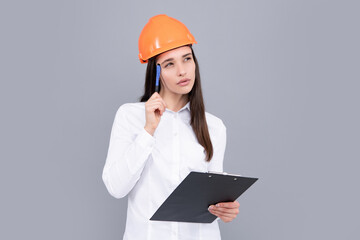 Thinking architect worker, female constructor engineer. Young beautiful architect woman in hard hat helmet hold clipboard over gray background. Female builder in helmet for advertising sign
