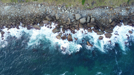 Aerial drone shot of waves hitting rock coastline with grass white and blue waves