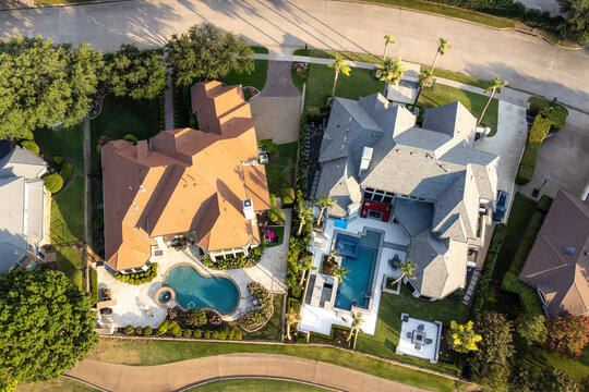 Aerial drone view of luxury mansions with swimming pools surrounded by green grass and trees in the summertime.