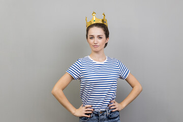 Portrait of woman wearing striped T-shirt and golden crown, looking at camera with confident facial expression, keeps hands on hips. Indoor studio shot isolated on gray background.