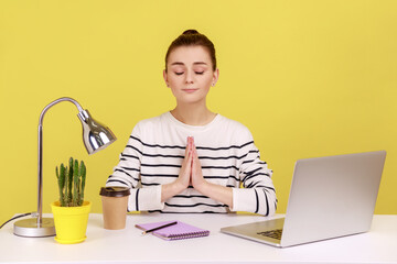 Open minded woman office worker in striped shirt pressing hands together meditating at workplace,...