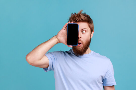 Portrait of bearded man covering eye with cellphone, hiding half of face, using gadget with wow shocked expression, trying to look at display. Indoor studio shot isolated on blue background.