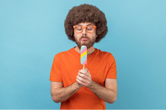 Portrait of man with Afro hairstyle wearing orange T-shirt and funny eyeglasses, holding ice cream, feels hungry, wants to lick dessert. Indoor studio shot isolated on blue background.