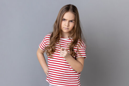 Portrait of little girl wearing striped T-shirt pointing finger at camera and looking with dissatisfied suspicious expression, warning about troubles. Indoor studio shot isolated on gray background.