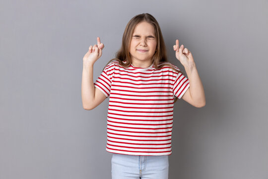 Portrait of little girl wearing striped T-shirt holding crossed fingers and wishing for good luck, having hopeful look, hoping for victory. Indoor studio shot isolated on gray background.