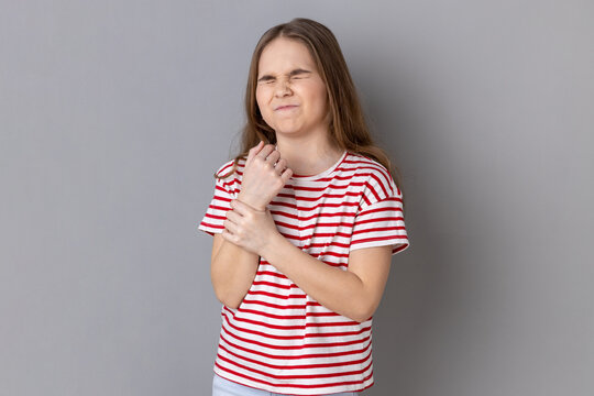 Portrait of sick little girl wearing striped T-shirt standing with grimace of pain, massaging sore wrist, suffering hand injury or sprain. Indoor studio shot isolated on gray background.