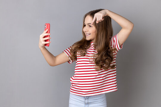 Little girl wearing striped T-shirt talking on video call and pointing down to subscribe, having online conversation on mobile phone, taking selfie. Indoor studio shot isolated on gray background.