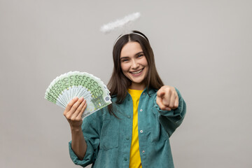 Positive angelic young woman with halo above head holding euro banknotes and pointing to camera, choosing lottery winner, encouraging to earn big money. Indoor studio shot isolated on gray background.