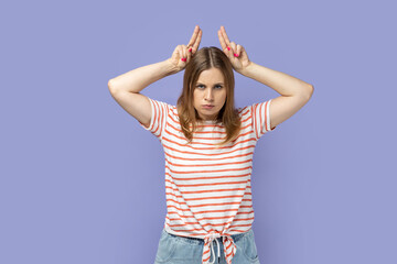 Portrait of angry bossy blond woman wearing striped T-shirt standing and showing horns, being anger...