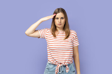 Responsible patriotic blond woman wearing striped T-shirt following discipline, saluting to commander with hand near temple and listening order. Indoor studio shot isolated on purple background.