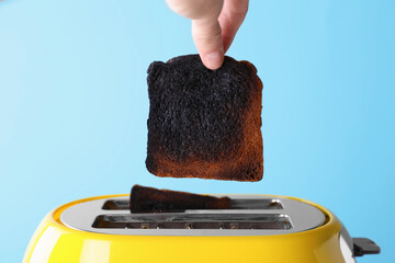 Woman taking off burnt bread from toaster against light blue background, closeup