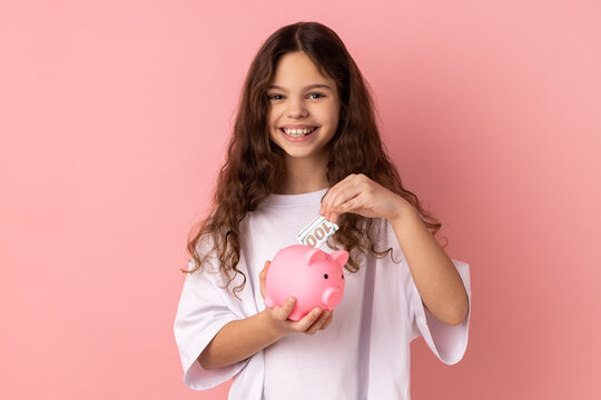 Portrait of delighted smiling little girl wearing white T-shirt standing looking at camera, putting dollar banknote into piggy bank. Indoor studio shot isolated on pink background.