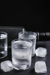Bottle of vodka and shot glasses with ice on table against black background