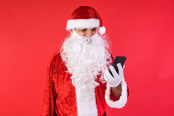 Man dressed as Santa Claus, consulting his mobile phone, on red background. Christmas, celebration, gifts, consumerism and happiness concept.