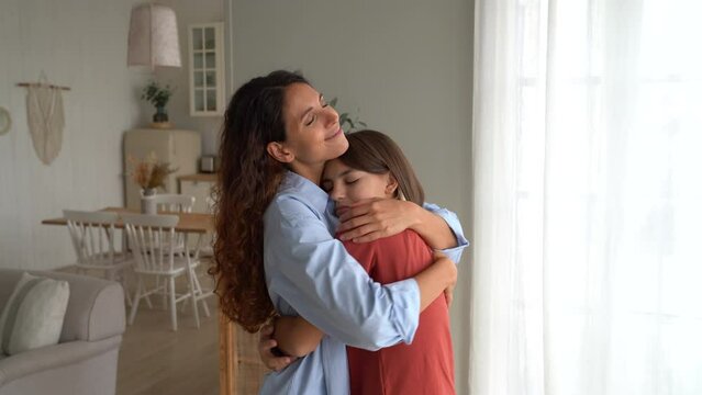 Loving teenage daughter tender hug mother woman at home and look at camera. Smiling mom and little girl embrace, reconcile after quarrel. Mother female show love have sweet moment together with baby.