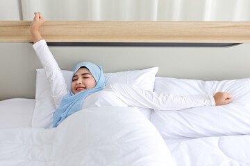 Beautiful asian woman wearing white muslim sleepwear lying on bed, stretching her arms after...